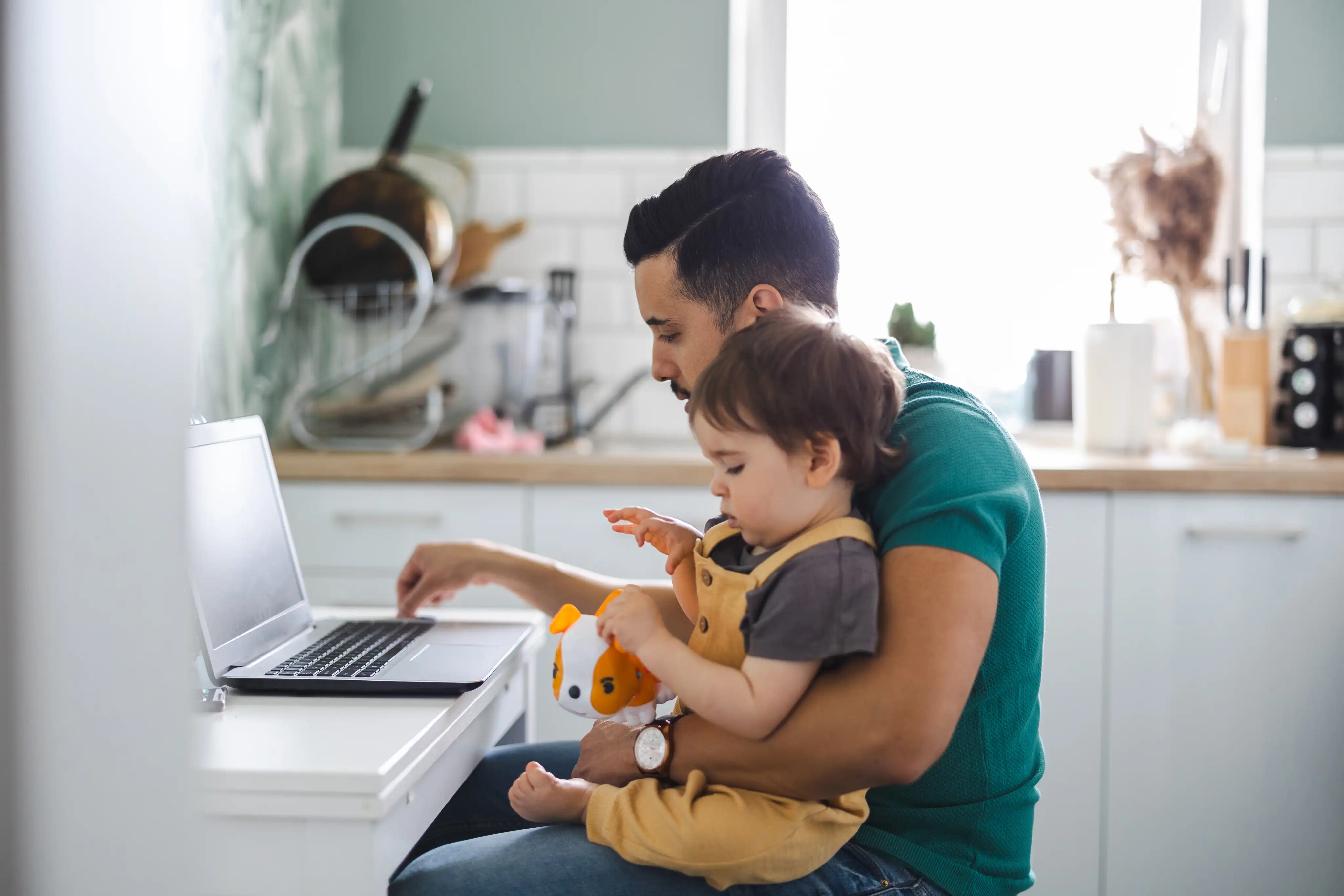 A dad and young son sitting in front of an open laptop in a kitchen.