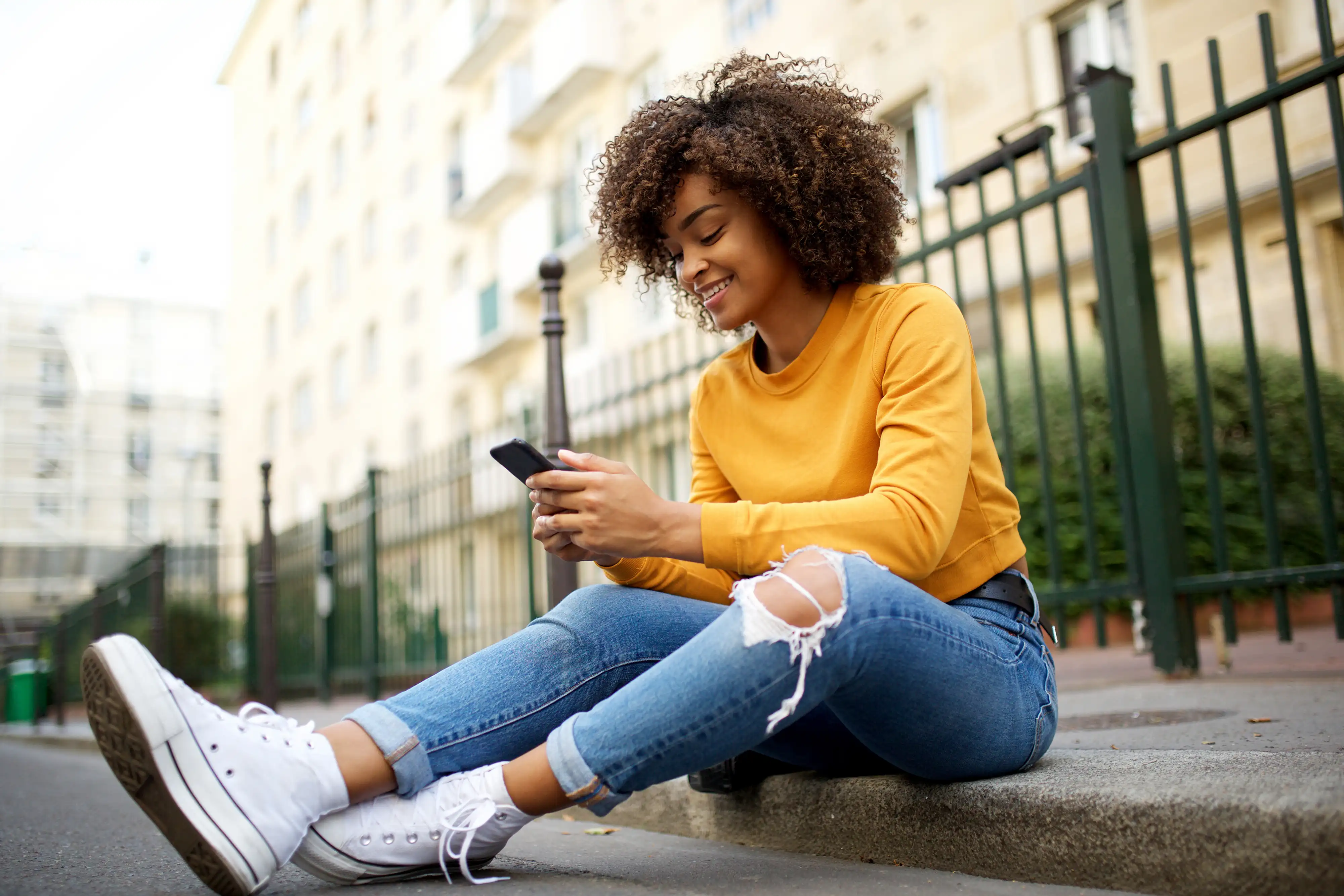 A happy woman sitting on a sidewalk looking at her phone.